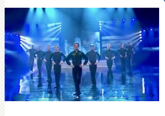 9 men line up to dance but notice the man in front when a woman with drums enters the stage